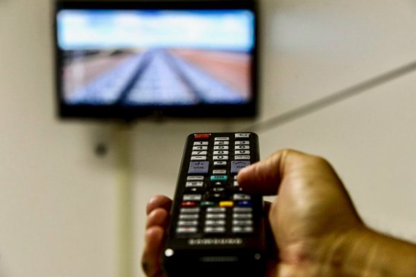 Pessoa usando controle remoto, apontando para um aparelho de TV, fixado na parede