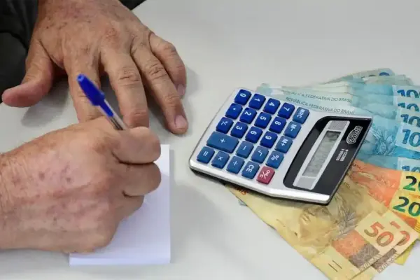 Pessoa usando calculadora para fazer contas. Notas de dinheiro também aparecem na imagem